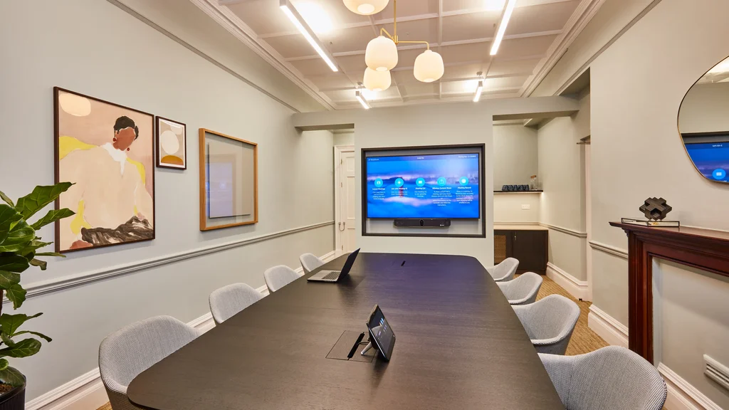 Meeting room area with large table and four chairs, with AV equipment including monitor and speakers for conference calls.