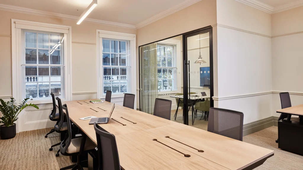 Office space with large communal desk area with six office chairs. Executive office behind communal desks.