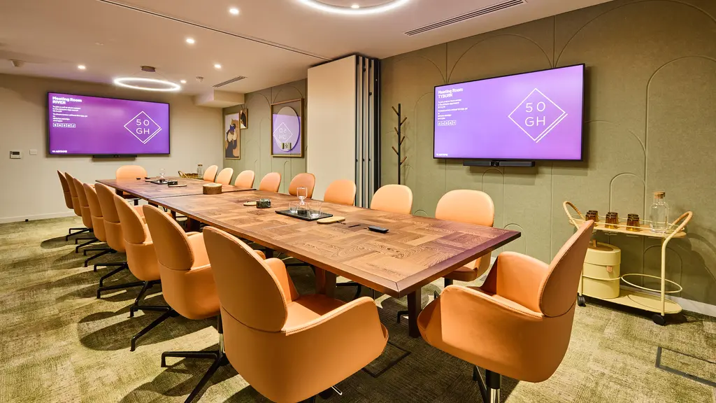 A meeting room with a long table surrounded by chairs and with two screens